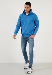 Felpa in pile blu con colletto a mezza zip, abbinata a jeans in denim chiaro e scarpe da ginnastica bianche sportive con dettagli blu e rossi.