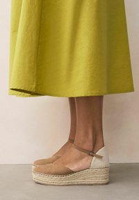 Le scarpe con zeppa in suede marrone presentano una suola in juta intrecciata e un accento in rete beige sul tallone, con un cinturino alla caviglia regolabile.