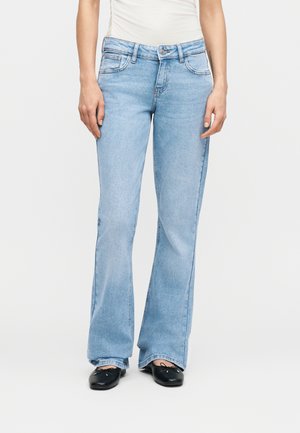 Woman wearing light blue high-waist flare jeans paired with black ballet flats, standing against a plain white background.