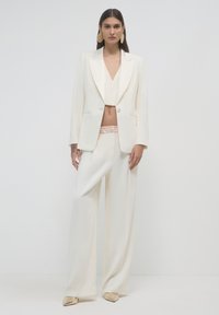 Blazer bianco sartoriale con accenti in satin, abbinato a pantaloni a gamba larga. Tessuto morbido, top a croppato con scollatura a V e tacchi metallici a punta.