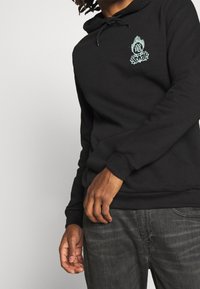 Czarny hoodie z kapturem i kieszenią z przodu, z grafikiem w odcieniu turkusu na klatce piersiowej, wykonany z miękkiego, teksturowanego materiału.