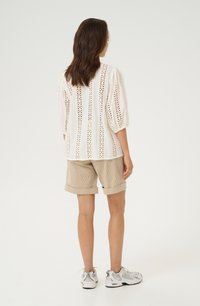 Blusa de algodón blanca con recortes decorativos, mangas abullonadas; combinada con pantalones cortos de pana beige. Modelo lleva zapatillas deportivas. Fondo simple.
