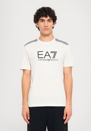 Light cream t-shirt with black logo "EA7 EMPORIO ARMANI" at the center. Features black and white striped accents on shoulders. Short sleeves.