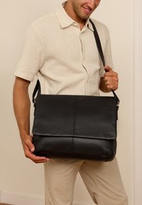 Černá kožená taška přes rameno s klopou, s texturovaným povrchem a nastavitelným popruhem, kterou drží osoba v béžové košili.