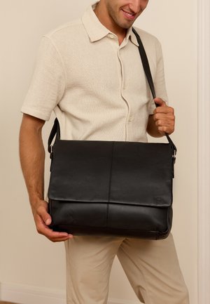 Bolso mensajero de cuero negro con cierre de solapa, que presenta una superficie texturizada y una correa ajustable, sostenido por una persona que lleva una camisa beige.