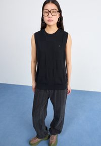 Top nero senza maniche in maglia con un motivo a griglia testurizzato, abbinato a pantaloni larghi a righe scure e sneakers marrone chiaro.