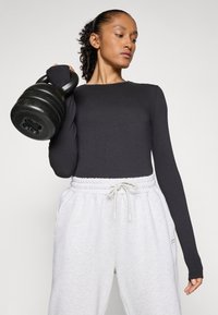 Frau in schwarzem Langarmoberteil und weißen Jogginghosen, die eine große schwarze Kettlebell auf ihrer Schulter hebt, vor einem schlichten Hintergrund.