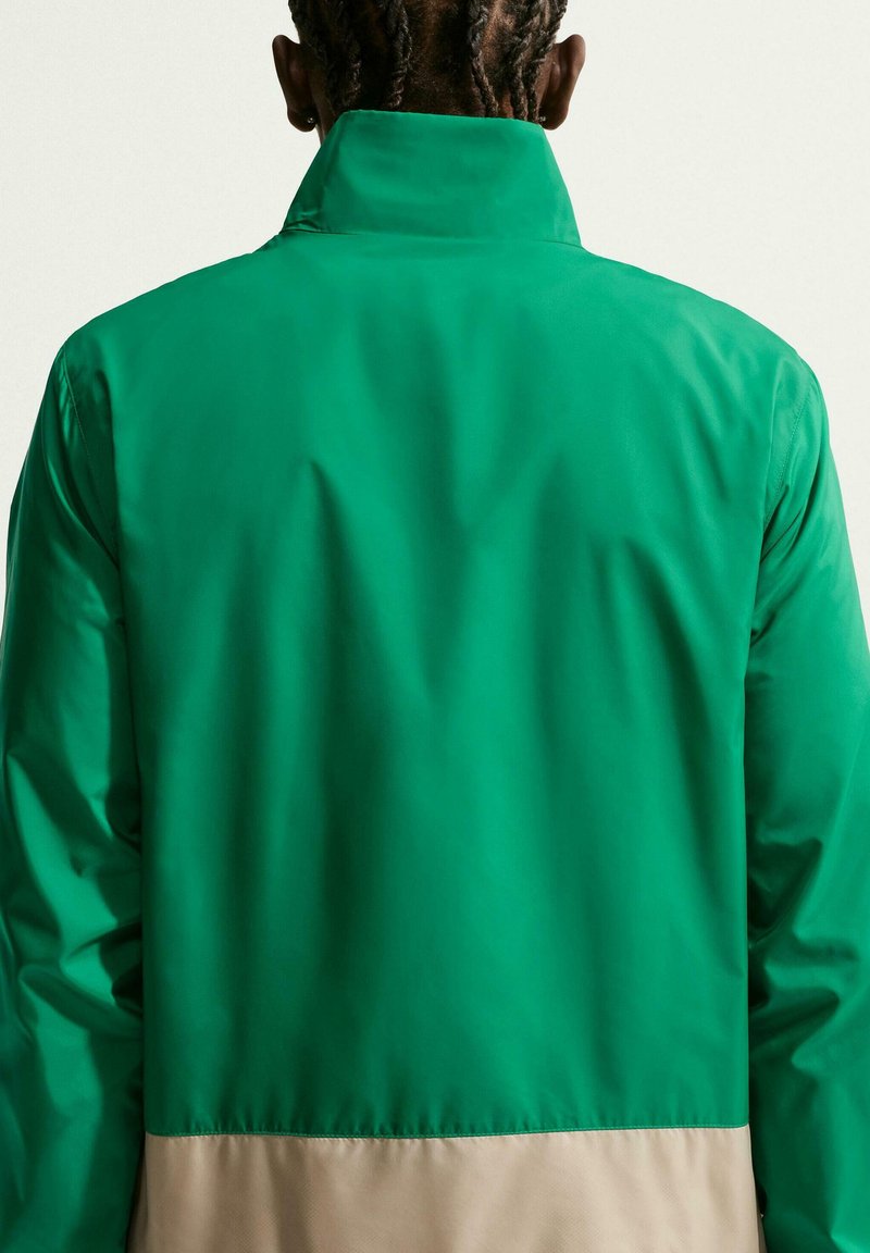 Person mit geflochtenem Haar von hinten gesehen, trägt eine grüne Jacke mit beigem Saum vor hellem Hintergrund.