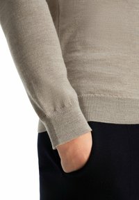 Beiger Wollpullover mit Rundhalsausschnitt und gerippten Bündchen, kombiniert mit dunklen Hosen. Weiche Textur, taillierte Passform und minimale Nahtdetails.