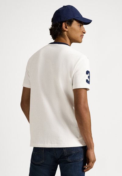 Λευκό t-shirt με κοντά μανίκια, navy γιακά και αριθμό 3 στο αριστερό μανίκι. Φορεμένο με σκουρόχρωμα τζιν και navy καπέλο. Λεία υφή.