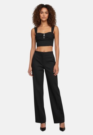 Donna con capelli ricci che indossa un top corto nero con bottoni e pantaloni neri a gamba larga e vita alta con tacchi neri, in piedi.