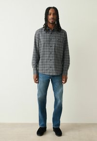 Camicia a quadri grigia con bottoni, maniche lunghe, abbinata a jeans blu e scarpe nere. La camicia presenta una tasca frontale e una vestibilità rilassata.