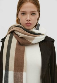 Sciarpa a quadri beige, marrone e grigia realizzata in tessuto morbido, avvolta attorno al collo con texture visibile e contrasto di colore rispetto a una maglietta bianca.