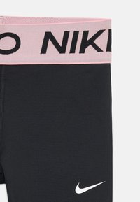 Čierne športové legíny s elastickým pásom v svetloružovej farbe, na ktorom je nápis "NIKE" čiernym písmom. Na nohe je viditeľný biely logotyp Nike.