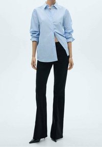 Lichtblauw gestreept overhemd met opgerolde mouwen, gecombineerd met zwarte uitlopende broek. Zwarte puntige hakken maken de outfit af.