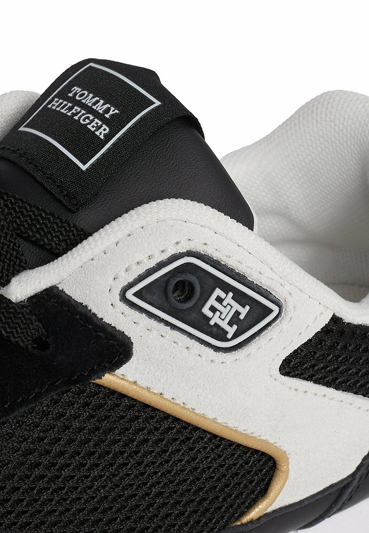Zapatilla deportiva en blanco y negro con parte superior de ante y malla, acentos metálicos en dorado y parche con logo en la lengüeta. Incluye cordones texturizados.
