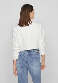 White cropped jacket with long, ruched sleeves, paired with high-waisted blue denim jeans featuring back pockets and a leather tag detail.