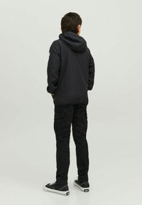 Černá bunda s kapucí a hladkou texturou, nošená přes černé cargo kalhoty. Oblečení je doplněno černými teniskami s bílými detaily.