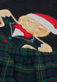 Temně modrý top s plyšovým perníkovým mužem, který nosí červenou vánoční čepici a motýlka, doplněný zelenou kostkovanou sukní s žlutými a červenými akcenty.