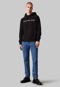 Sudadera con capucha negra y logo blanco "Calvin Klein Jeans", combinada con unos vaqueros azules y zapatos negros de cordones. Diseño simple y casual.