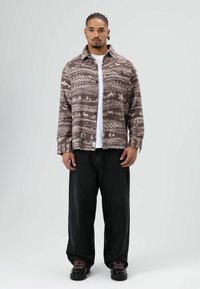 Bruine overhemd met geometrische patronen, knoopsluiting aan de voorkant en kraag, gecombineerd met zwarte wijde broekspijpen en donkere schoenen.