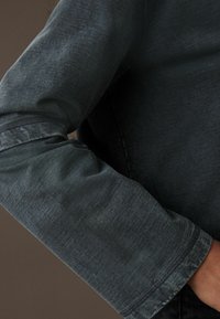 Mørkeblå denimærme med en let struktureret overflade. Har en manchet med stikninger og en afslappet pasform, der er velegnet til casual brug.