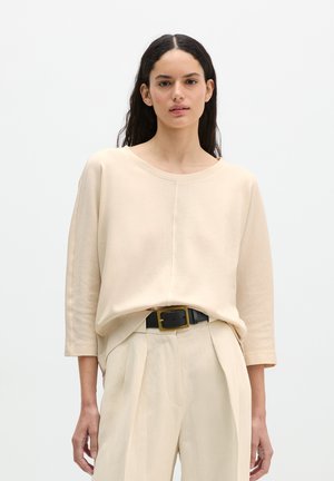 Vrouw met lang donker haar die een losse beige top met driekwart mouwen, beige geplooide broek en een zwarte riem met een gouden gesp draagt.