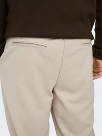 Beige textured pants with two back pockets featuring narrow openings, paired with a dark brown sweater. The fabric appears sturdy and woven.