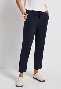 Navy blue tapered trousers with elastic waist and drawstring. Features side pockets and a smooth, lightweight fabric. Worn with white sneakers.