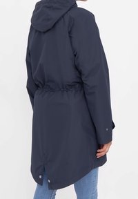 Marinblå vattentät jacka med huva, åtdragen midja och tryckknappsdetaljer. Har en längre bakre fåll och elastiska ärmslut.