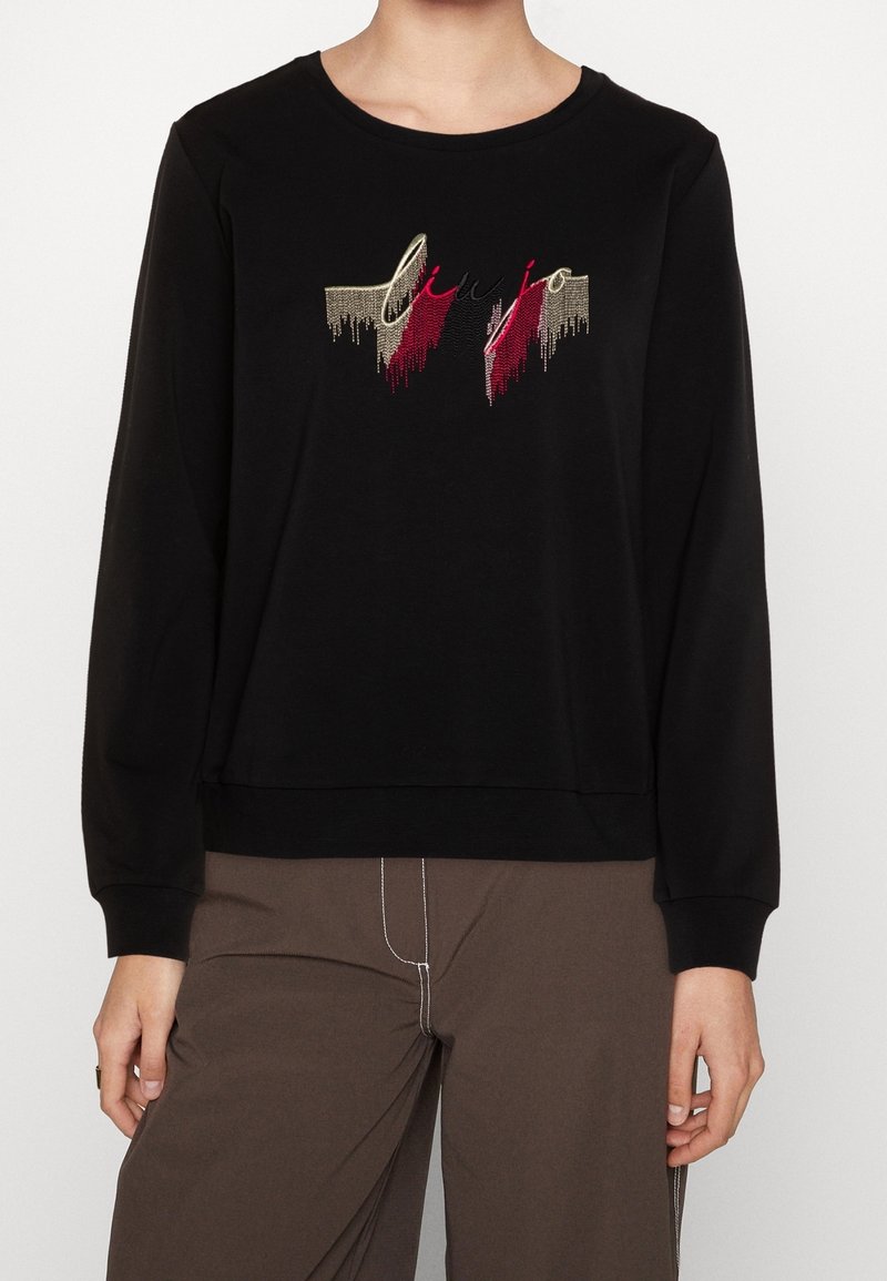 Person trägt ein schwarzes Langarmshirt mit einem roten und beigen abstrakten Design sowie braune Hosen mit weißer Naht.