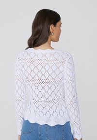 Biały sweter z dzianiny z wzorem w romby i falbaniastym dołem. Długie rękawy i teksturowany materiał zapewniają przewiewny, otwarty design.