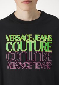 Svart t-shirt med djärv, flerfärgad text: "VERSACE JEANS COUTURE," med livliga gröna, gula och rosa bokstäver i olika orienteringar.