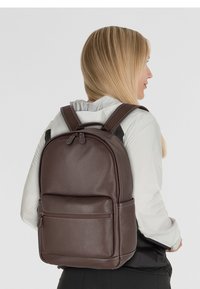 Sac à dos en cuir brun avec une surface texturée, doté de bretelles réglables, d'une poche zippée à l'avant et d'un design arrondi.