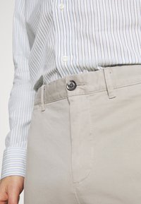 Světle béžové kalhoty s hladkým povrchem, vybavené zapínáním na knoflík a bočními kapsami, v kombinaci s modro-bílou pruhovanou košilí.