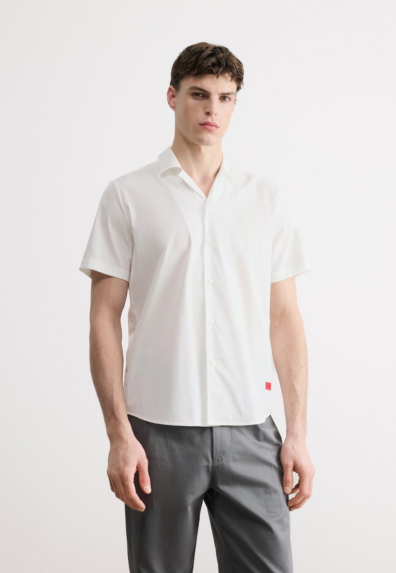 Νεαρός άνδρας που στέκεται φορώντας λευκό πουκάμισο με κοντά μανίκια και γκρι παντελόνι, μπροστά σε απλό ανοιχτό φόντο.