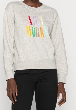 Person trägt ein hellgraues Sweatshirt mit der Aufschrift "ART WORKS" in bunten, hohen Buchstaben über der Brust, kombiniert mit einer schwarzen Hose.
