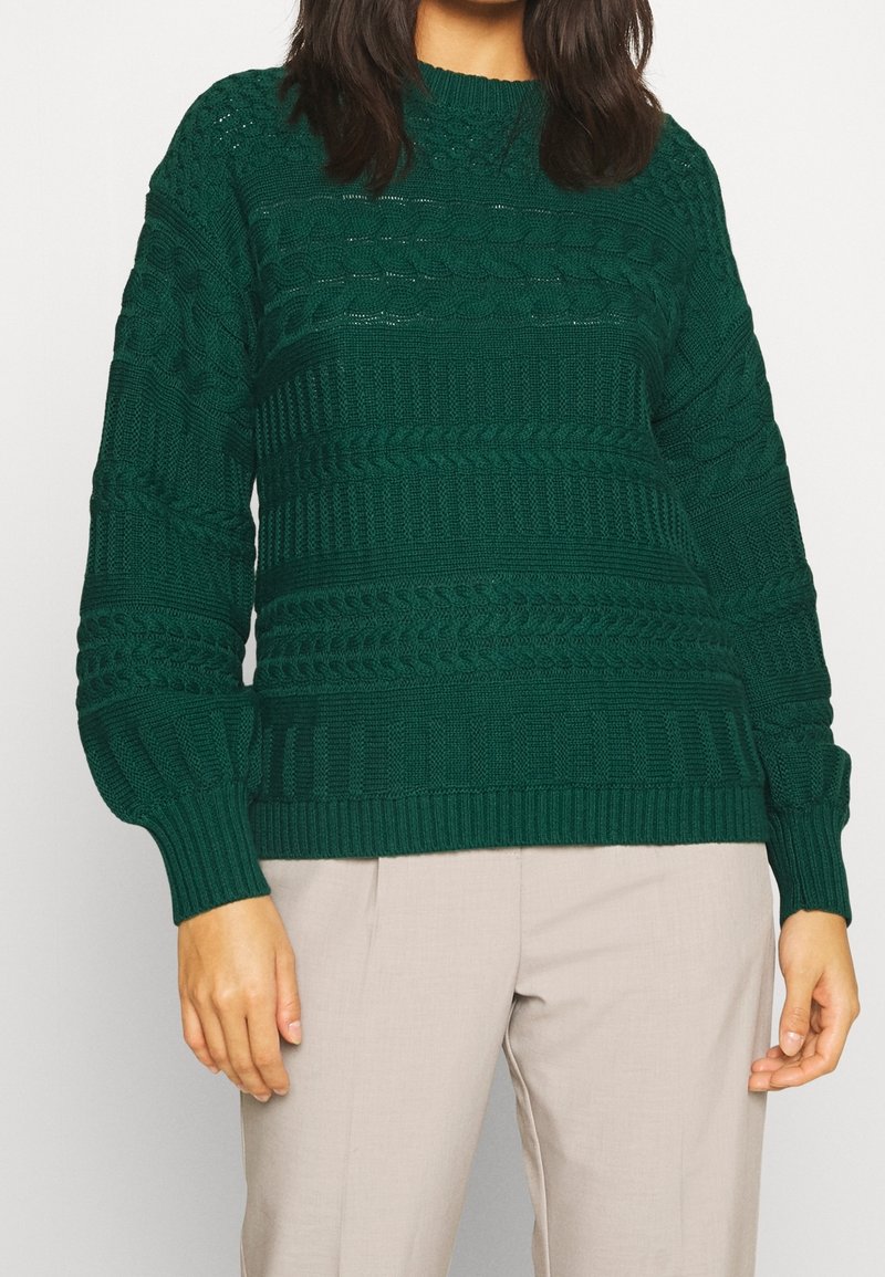 Pull vert en maille avec motifs texturés, poignets et ourlet côtelés. Associé à un pantalon beige clair, coupe décontractée.