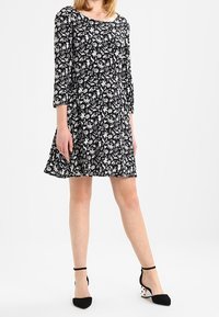 Robe noire à motifs floraux avec des manches trois-quarts et une jupe évasée. Présente des motifs de fleurs blanches. Associée à des chaussures à talons noires et blanches.