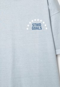 Lysblå skjorte med mørkeblåt tekst "STWD GOALS" under en bue af hvide stjerner på den øverste venstre brystområde.