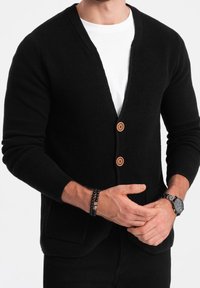 Homme portant un cardigan en maille noire avec deux boutons marron par-dessus une chemise blanche, avec un bracelet noir à perles et une montre-bracelet en métal.