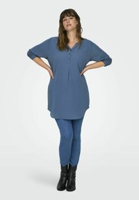 Túnica azul con mangas tres cuartos, escote henley abotonado, tela suave, combinada con leggings azul oscuro y botas negras hasta el tobillo.