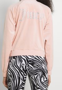 Rosa velourjacka med rhinestone-texten "Juicy" på ryggen. Har långa ärmar och ribbad fåll. Matchas med svartvita byxor med abstrakt mönster.