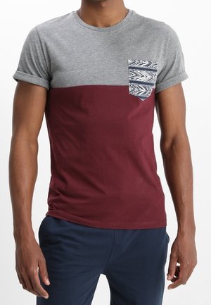 Mand iført en kortærmet t-shirt med grå øverste halvdel, mørkerød nederste halvdel og et mønstret brystlomme, kombineret med mørke bukser.