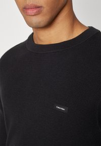 Maglione nero a maniche lunghe con scollatura rotonda, realizzato in tessuto tessuto con un'etichetta Calvin Klein cucita sul petto sinistro. Design semplice e minimalista.