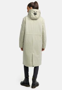 Lichtgroene lange jas met een capuchon, voorzien van een rugventilatie en verstelbare trekkoorden. Zwarte accenten en stevige knopen aan de zoom.