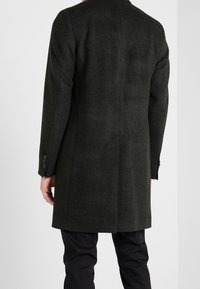 Abrigo de mezcla de lana verde oscuro con una forma ajustada, que presenta una abertura en la parte posterior y puños abotonados, llevado sobre pantalones negros.