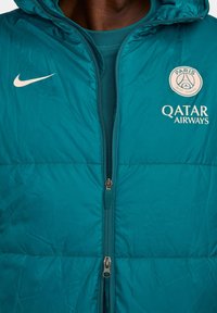 Jachetă puffer teal cu închidere cu fermoar, având logo-ul Nike și emblema Paris Saint-Germain, cu textură netedă și glugă.