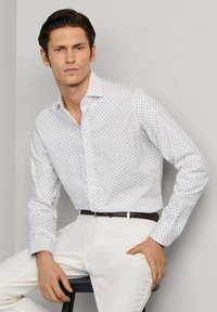 Muškarac obučen u bijelu, uzorkom ukrašenu košulju s dugmadima i bijele hlače, sjedi na crnoj stolici ispred običnog zida.