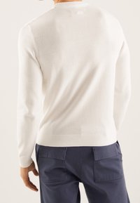 Helles cremefarbenes Strickpullover mit Rundhalsausschnitt, geripptem Saum und langen Ärmeln, kombiniert mit marineblauen Hosen mit Gesäßtaschen.